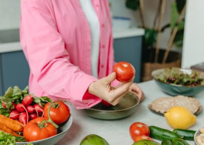 Person in pink overshirt holding a fresh tomato with various organic vegetables on a countertop.