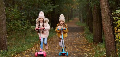 Two girls wearing beanies and winter jackets ride scooters on a leaf-strewn path in an autumn park.