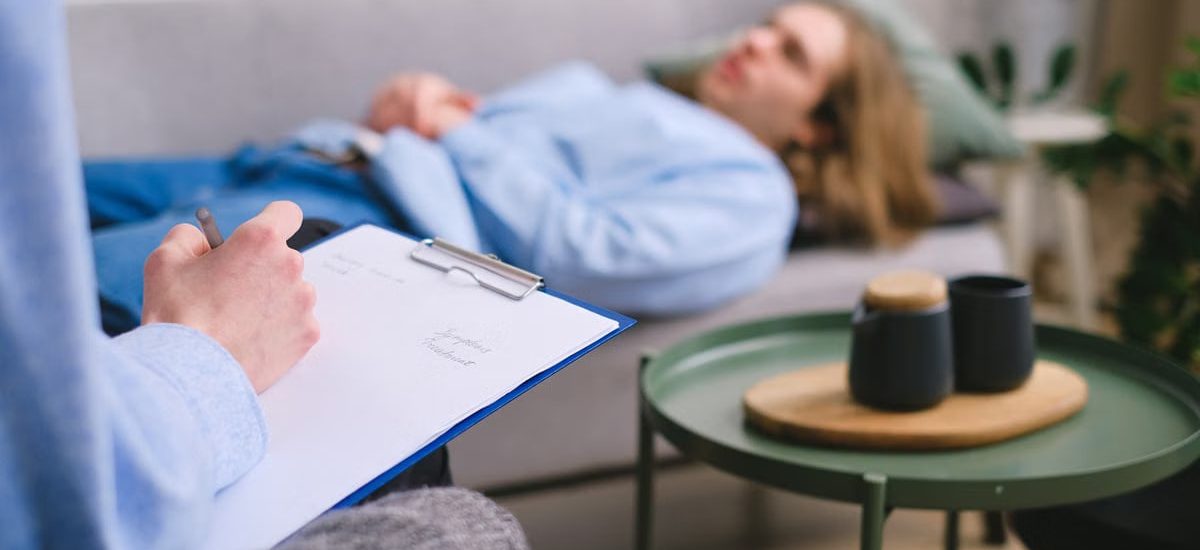 A therapist taking notes during a session with a patient lying on a sofa indoors.