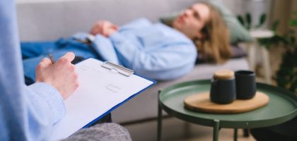 A therapist taking notes during a session with a patient lying on a sofa indoors.